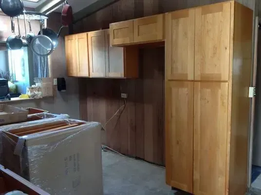 Kitchen cabinets, some installed, in natural wood color, with unfinished walls, near a window.