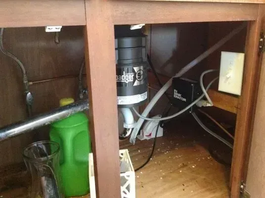 Under-sink cabinet with garbage disposal, pipes, electrical outlet, cleaning supplies, and debris.