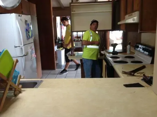 A kitchen with two men: one in the background, walking, the other near the stove wearing a safety vest.