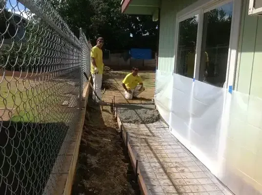 Two workers in yellow shirts pouring concrete along a building wall next to a fence.