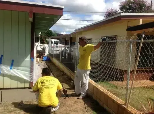 Two men in yellow shirts working on a building, one kneeling, other leaning on a fence.