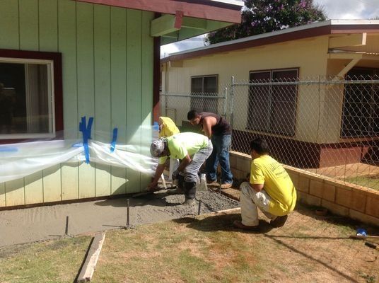Construction workers pouring concrete next to a light green building. They are wearing safety gear and working outdoors.