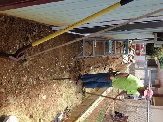 A man in a neon shirt digs soil near a building, next to a ladder and a long-handled tool.
