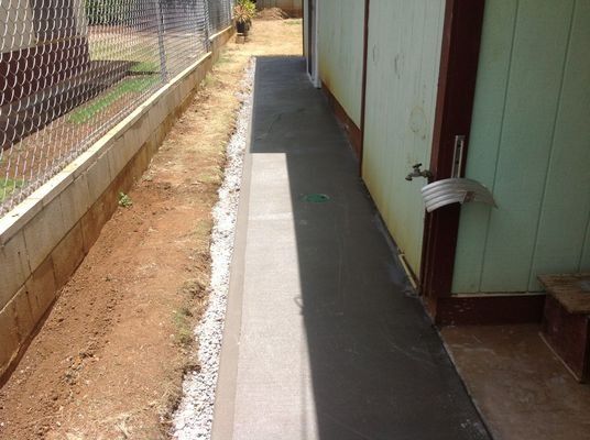 A concrete walkway next to a building and chain-link fence, with dirt on one side.