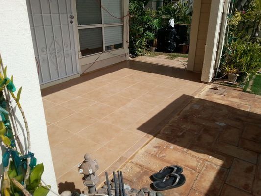 Tiled outdoor patio with a doorway, flip-flops, and garden. Brown tiles and sunlight.