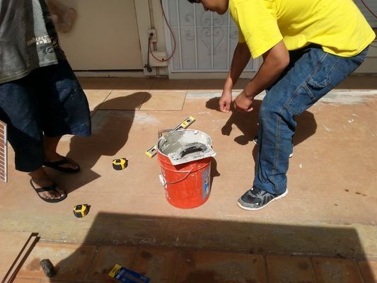 Two people working on a floor outside: one in yellow shirt, one in jeans, with tools and a bucket.