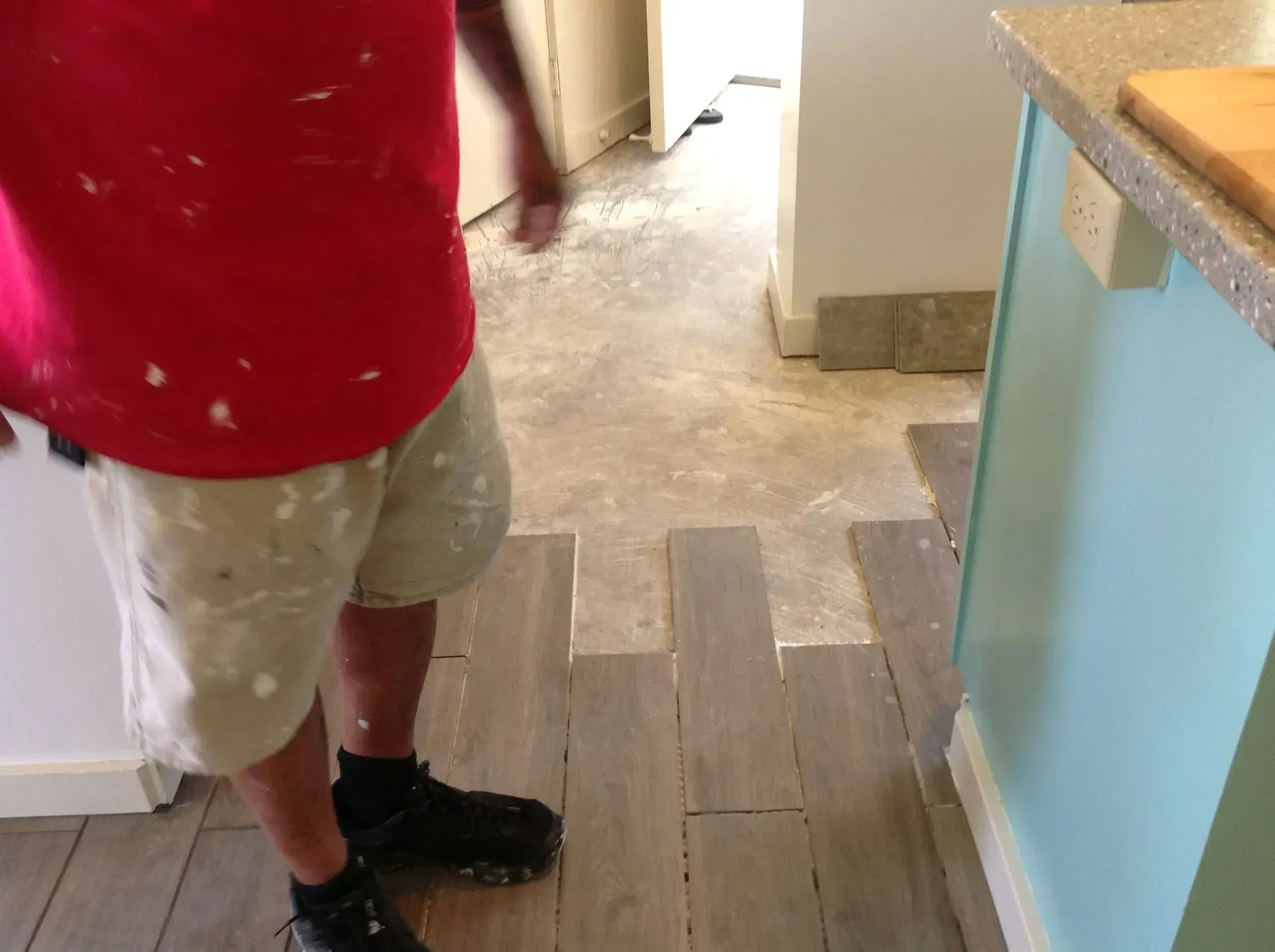 Person in red shirt and paint-splattered shorts installing wood-look tile flooring in a kitchen.