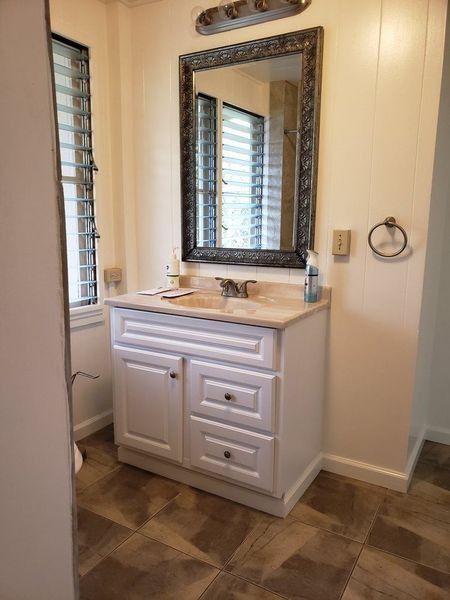 Bathroom with white vanity, framed mirror, and window with blinds. Brown floor and light beige walls.