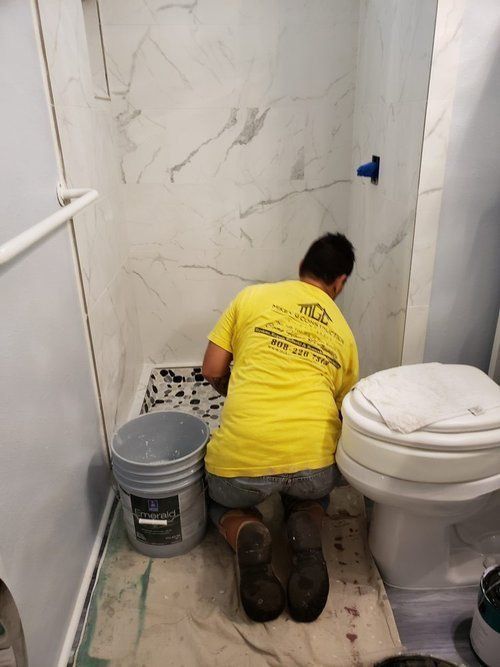 A person kneels in a bathroom, tiling the floor. They wear a yellow shirt and dark boots.