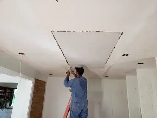 Person on a ladder cutting a square hole in a white ceiling with a drywall knife.