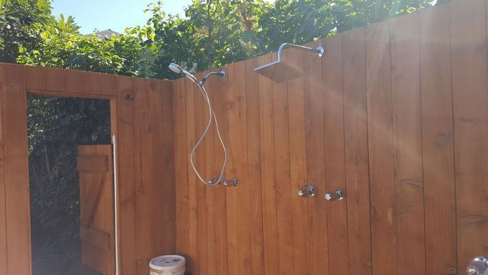 Outdoor wooden shower with silver fixtures, against a wooden wall; bright sunlight.