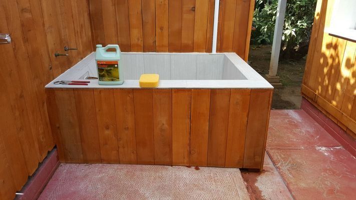 Wooden outdoor bathtub with white tiled interior, surrounded by wooden walls. A cleaner and sponge sit on the edge.