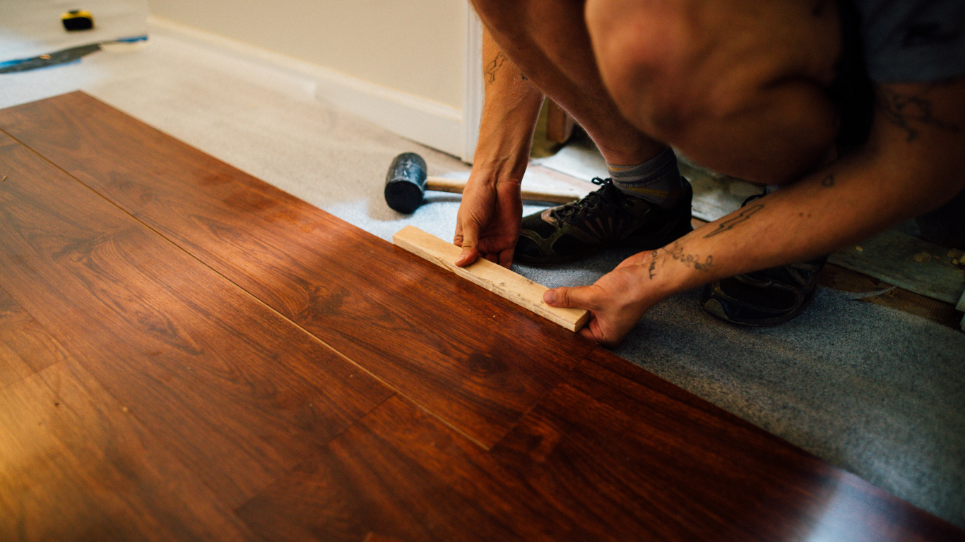 Person installing wood flooring, using a spacer. Dark brown planks, beige walls.