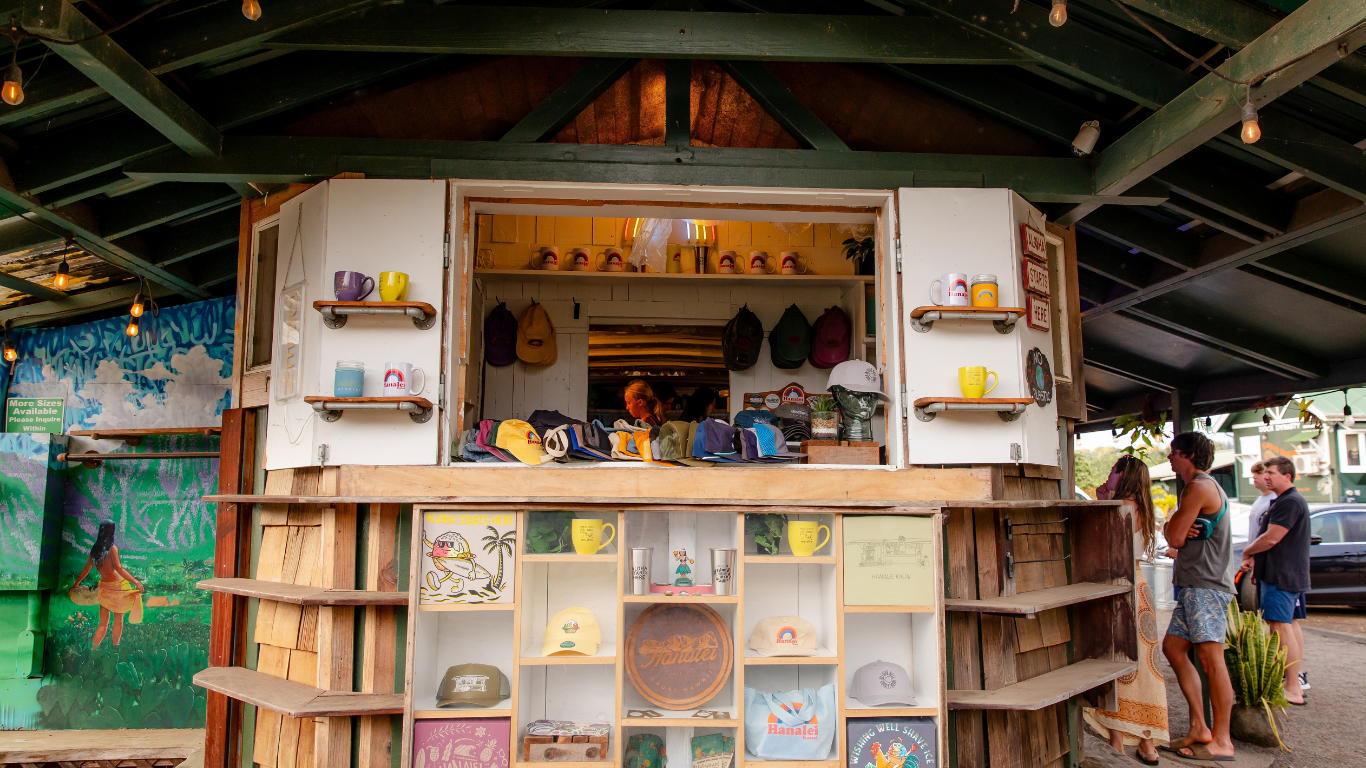 A small, outdoor shop with customers. White exterior, open window displays goods. Shelves are stocked, two people browsing.