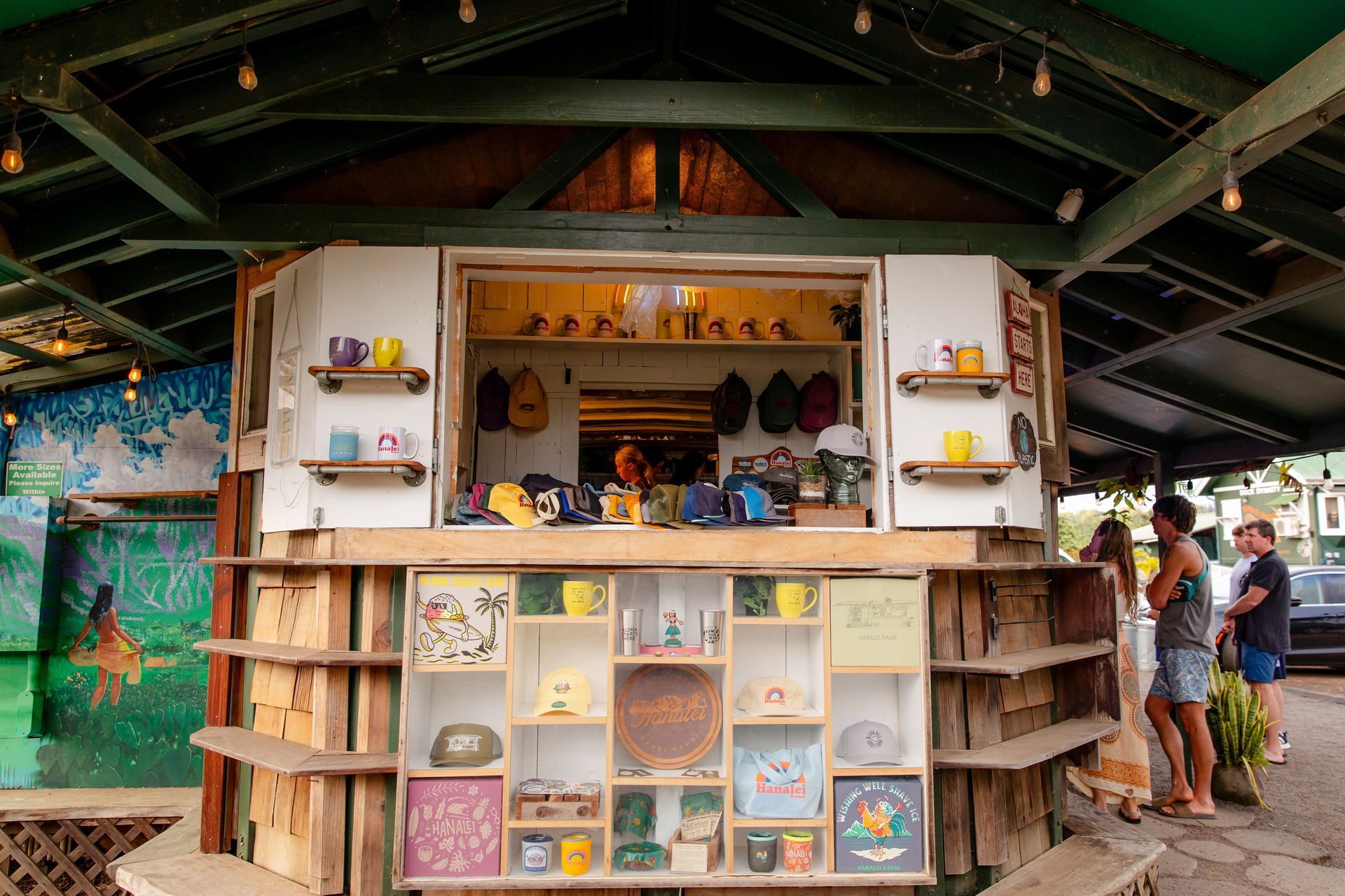 A colorful outdoor shop with customers. White and wood structure with merchandise displayed.