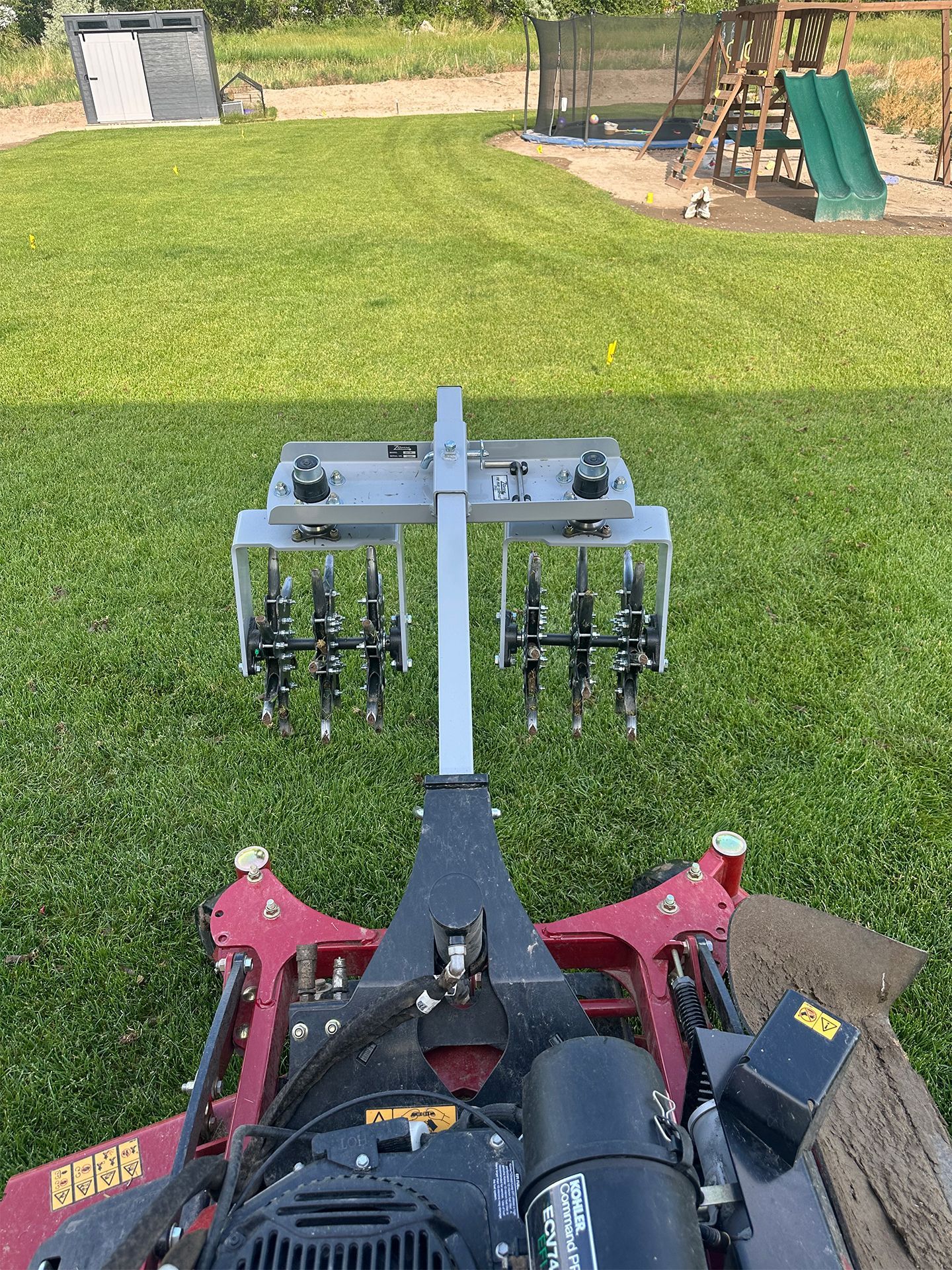 Lawn aerator attachment on riding mower. Metal spikes on a green lawn, background shows playground and shed.