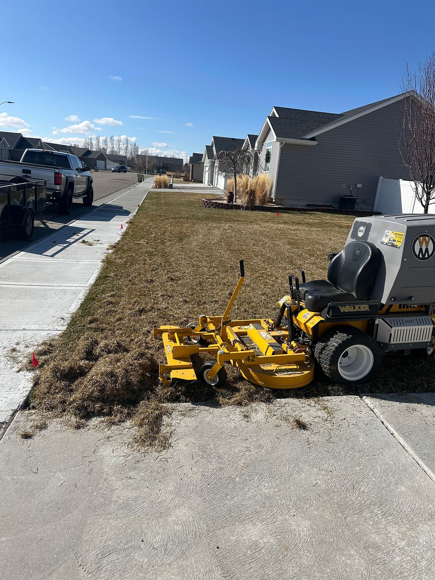 Yellow lawn mower mulching dried grass on a residential lawn, with houses and a truck in the background.