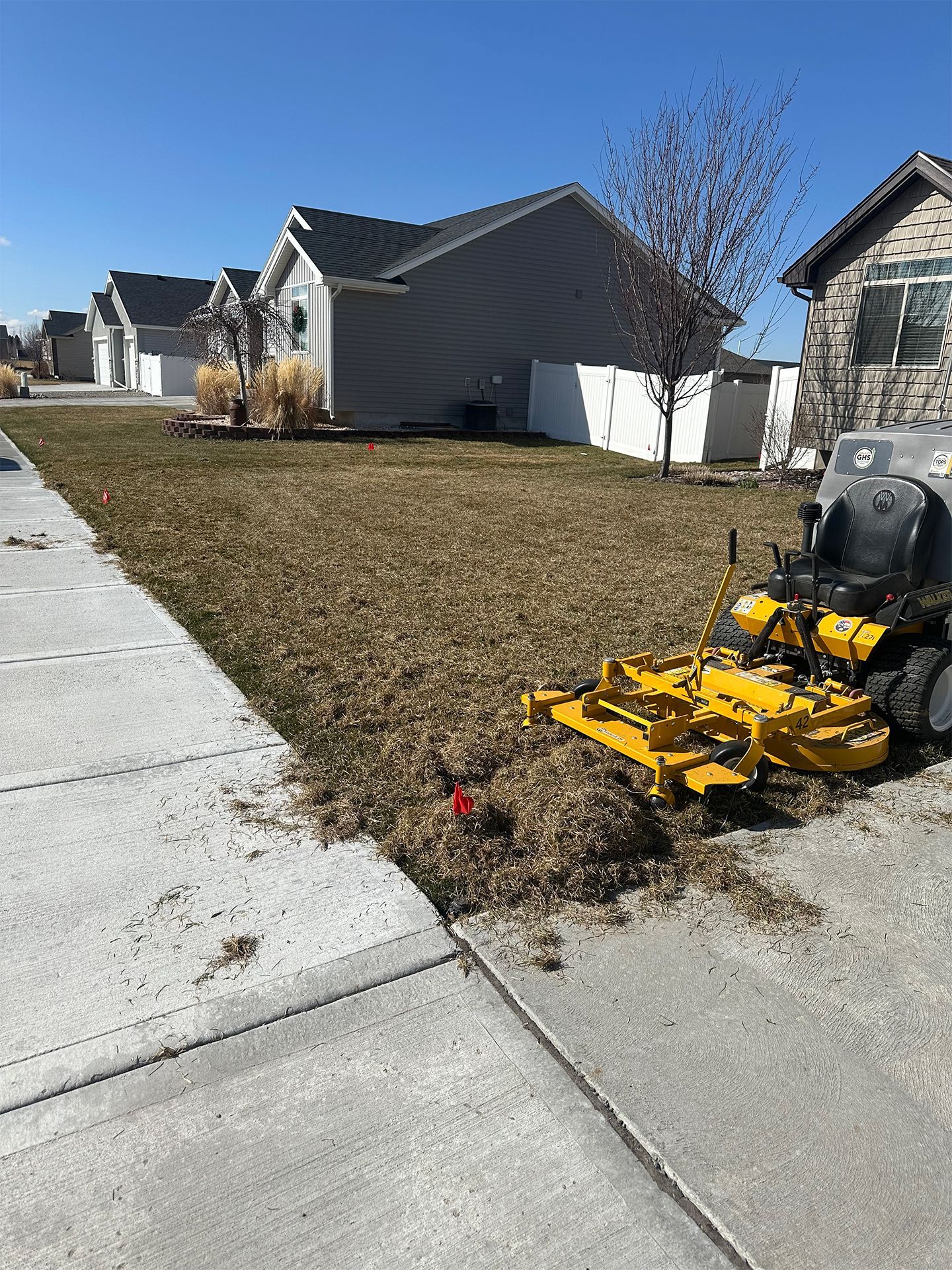 Yellow lawnmower on a residential lawn, next to a sidewalk. Brown grass and houses in the background.