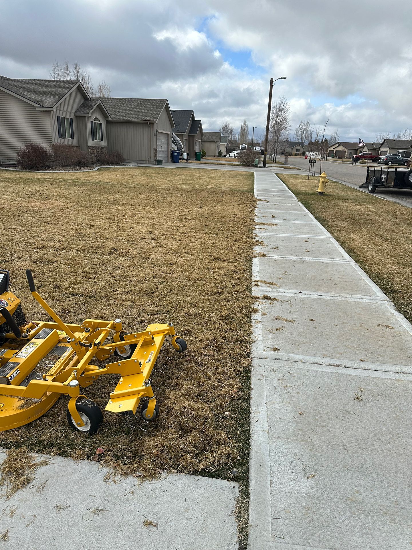 Lawn mower on brown grass next to a sidewalk in a residential neighborhood. Cloudy sky.