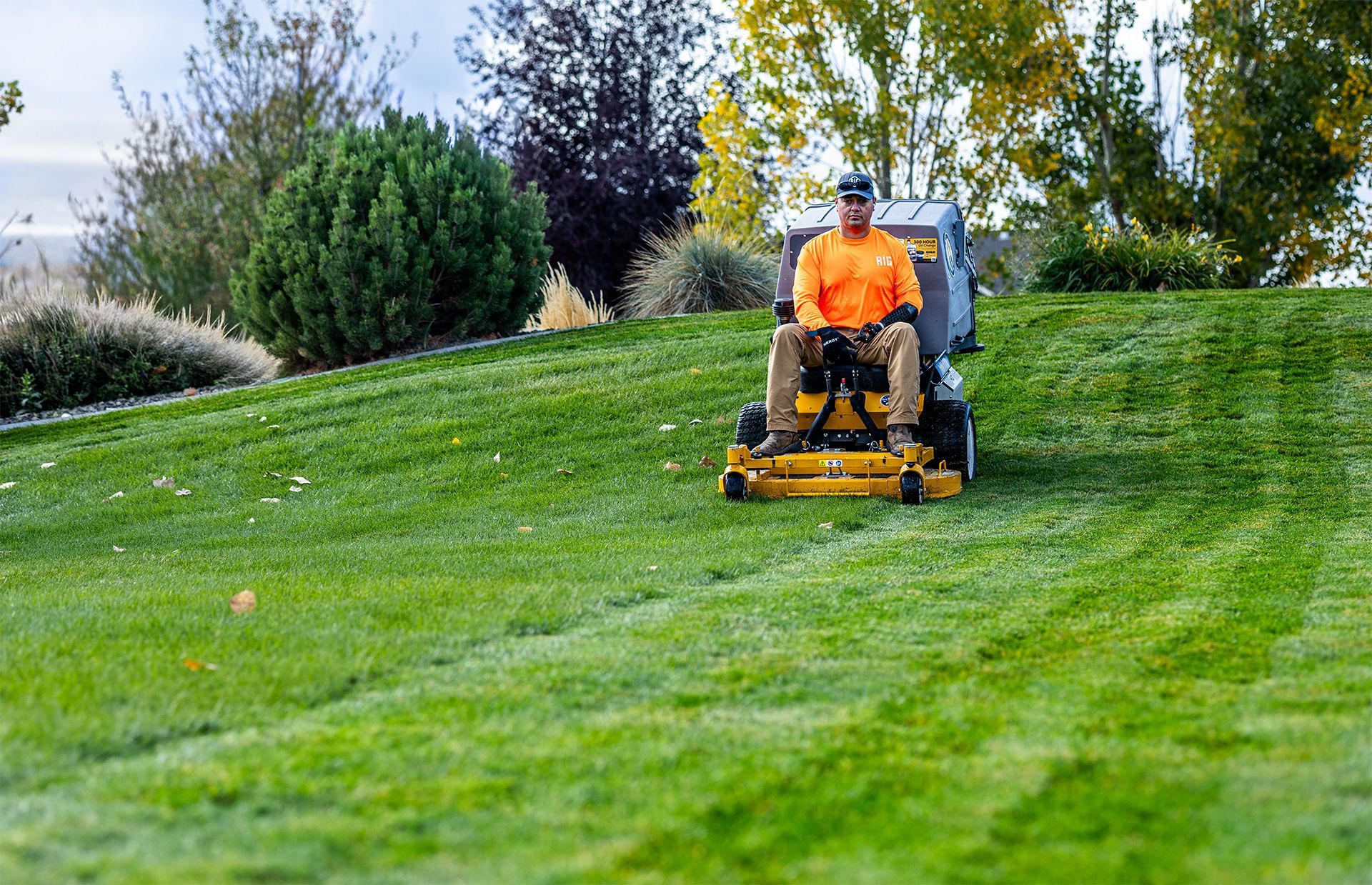 Person in orange shirt operating a riding lawnmower on a sloped, grassy yard.
