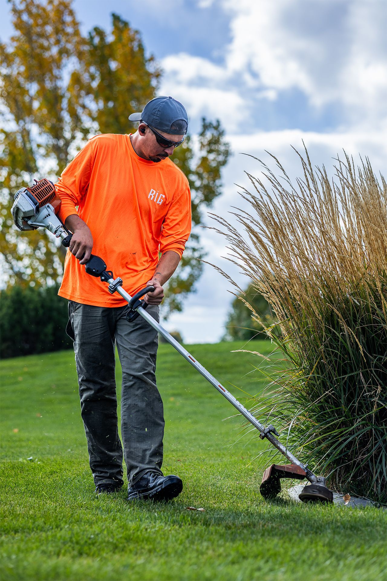 Man in orange shirt using a weed trimmer on a grassy lawn with a bush in the background.