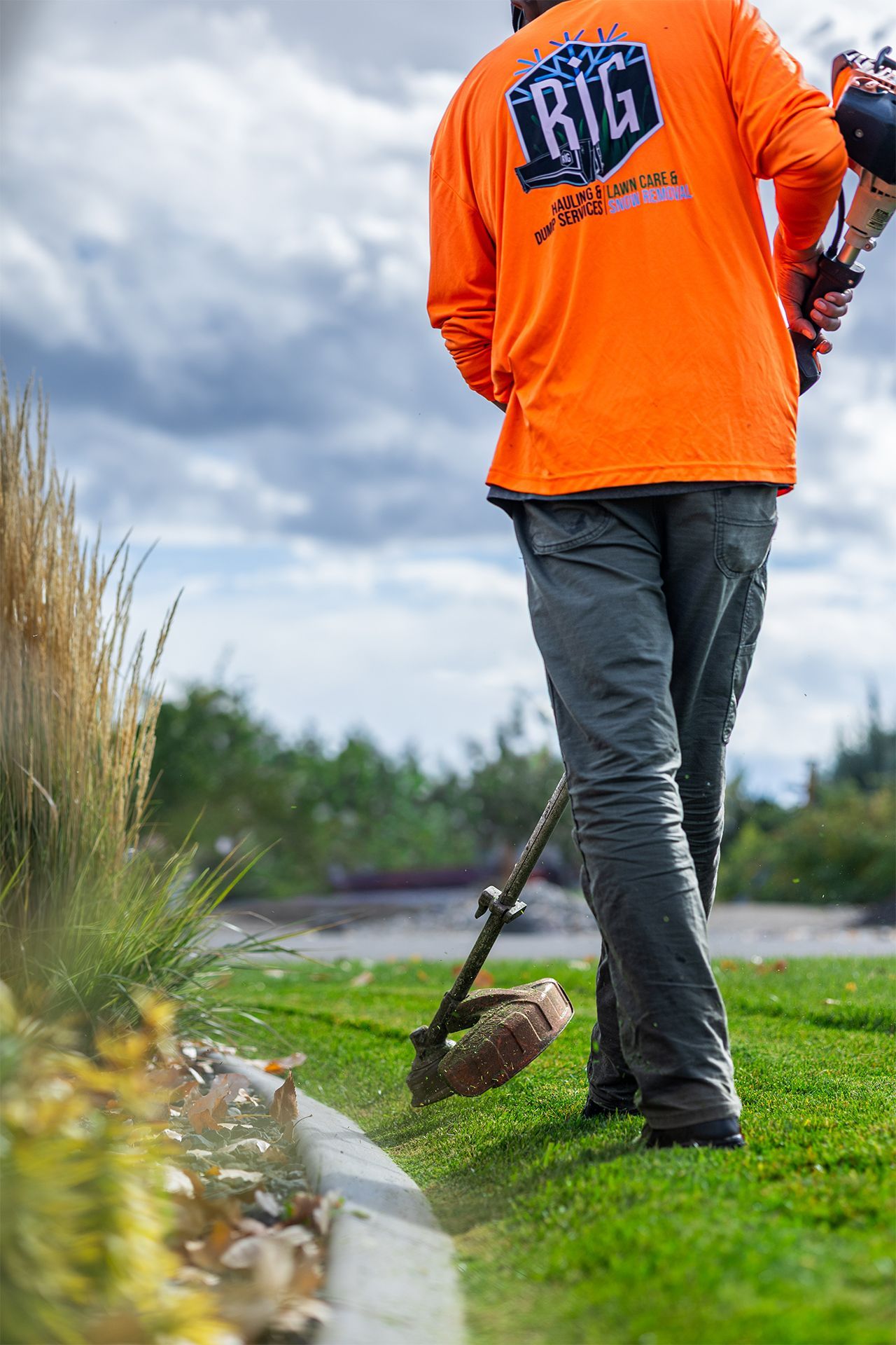 Man in orange shirt using a weed whacker near a curb and grass. Cloudy sky in the background.