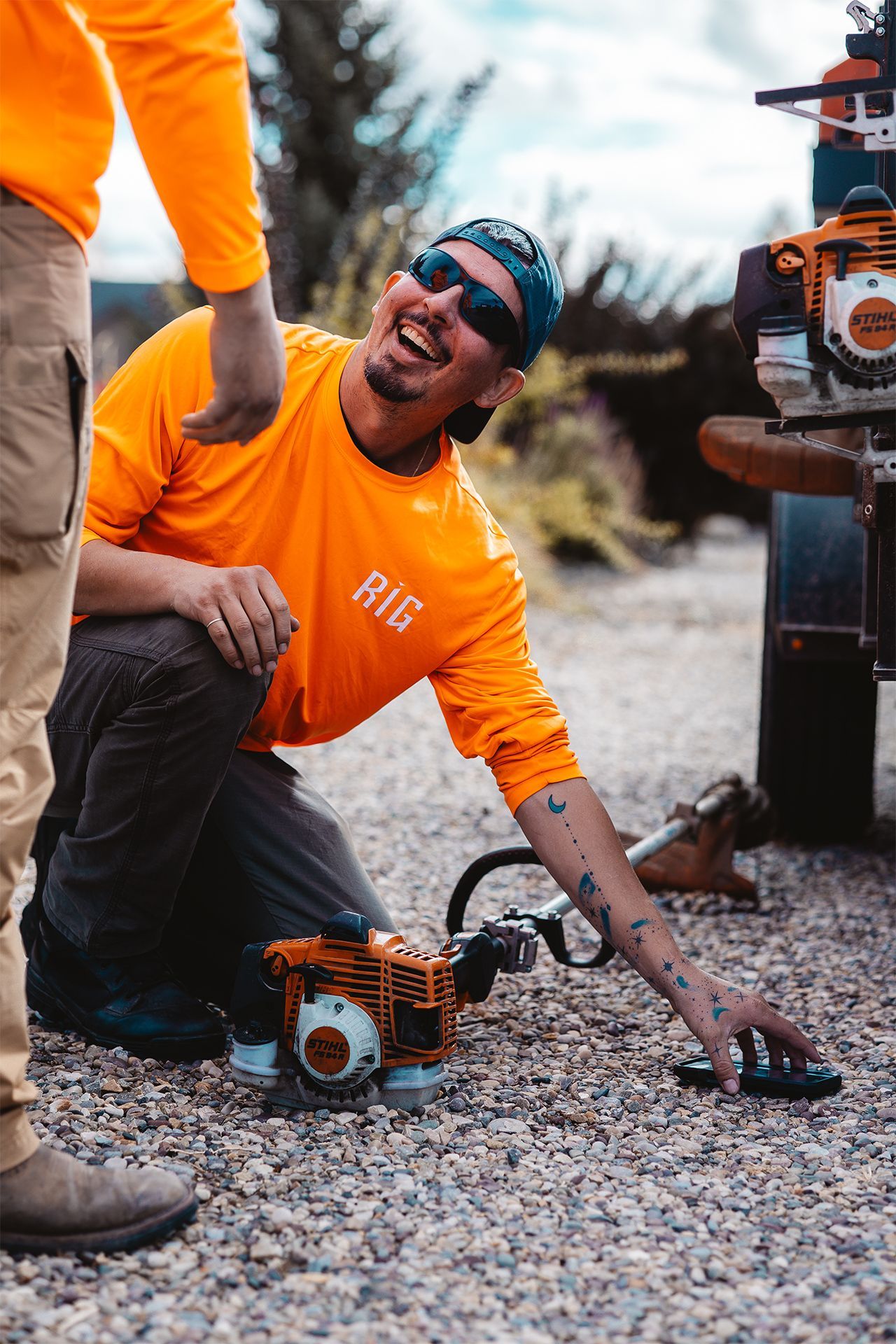 Man in orange shirt kneels with a weed trimmer; smiling. Another person nearby. Gravel ground, sunny outdoors.