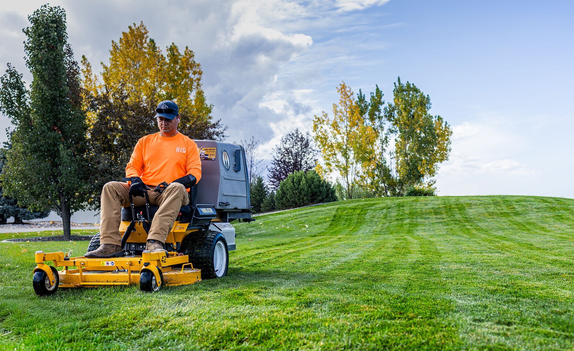 Man operating a yellow lawn mower on a grassy hill; blue sky and trees in the background.