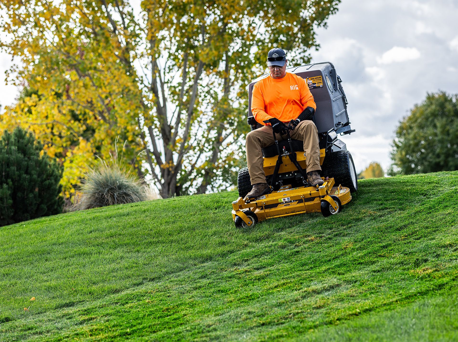 Person on a yellow riding lawnmower mowing grass on a green hill with fall foliage in the background.
