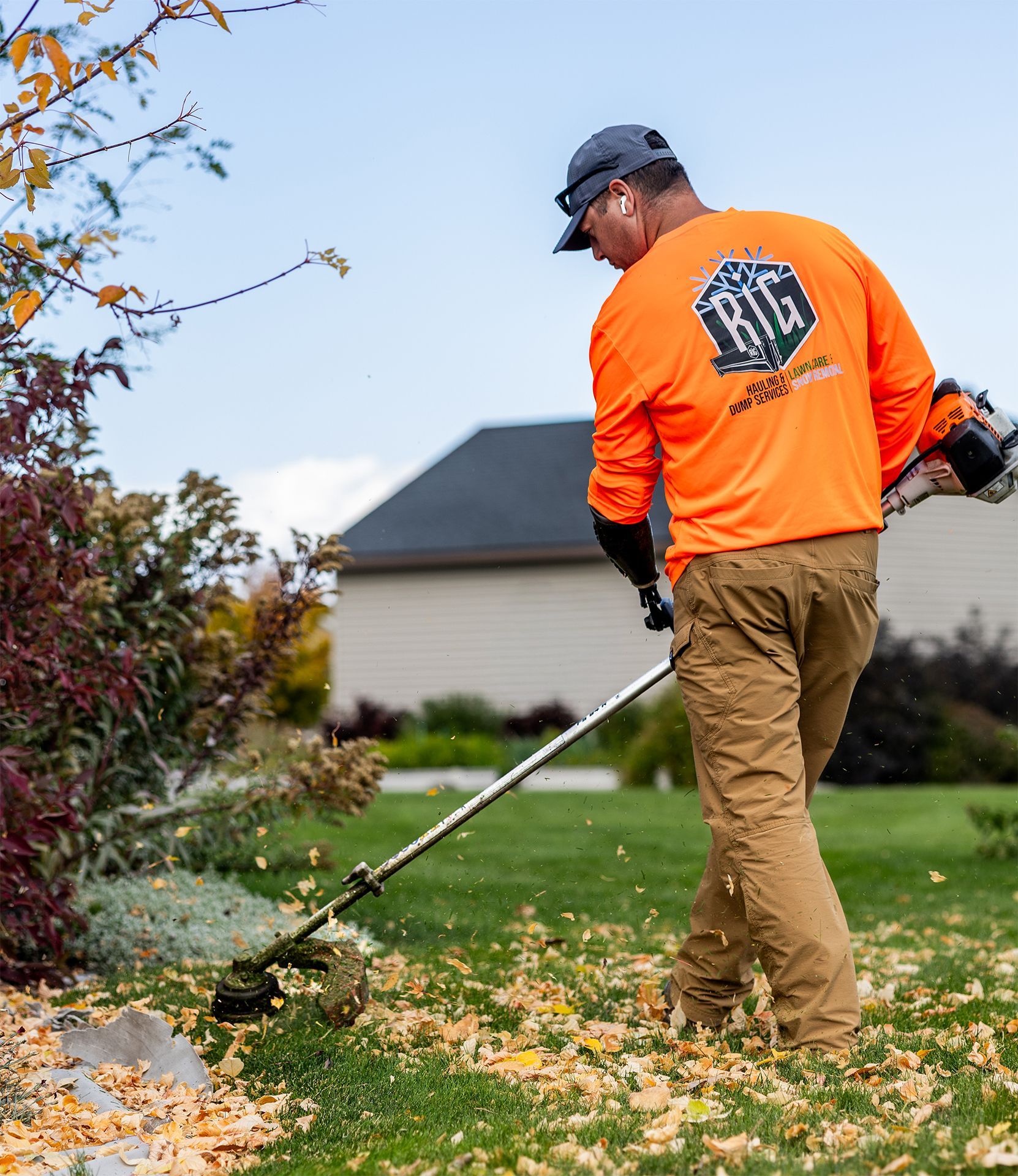 Man in orange shirt using a weed whacker on a lawn with fallen leaves.