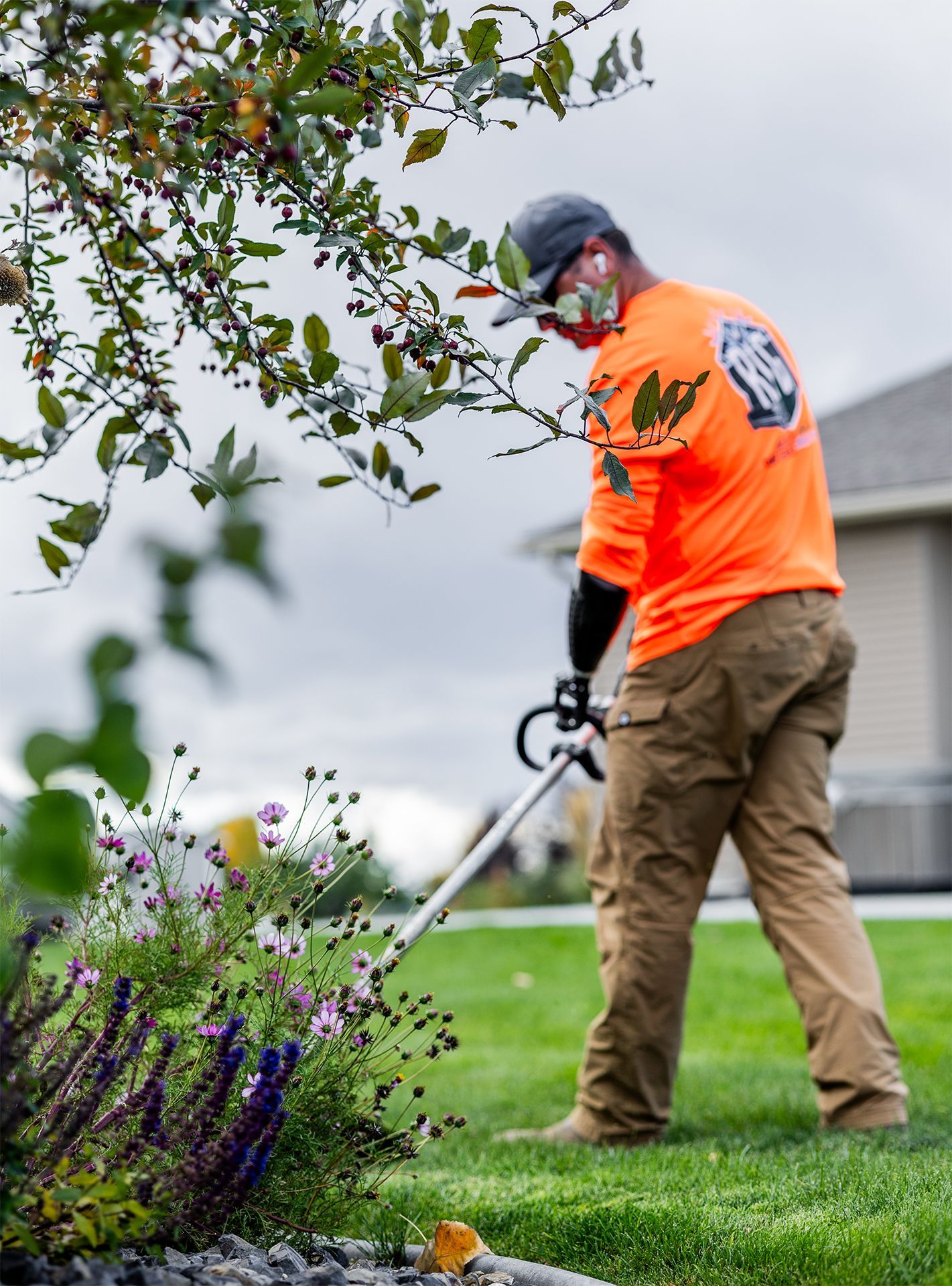 A person in orange shirt trims grass with a weed wacker near a flower bed and tree.