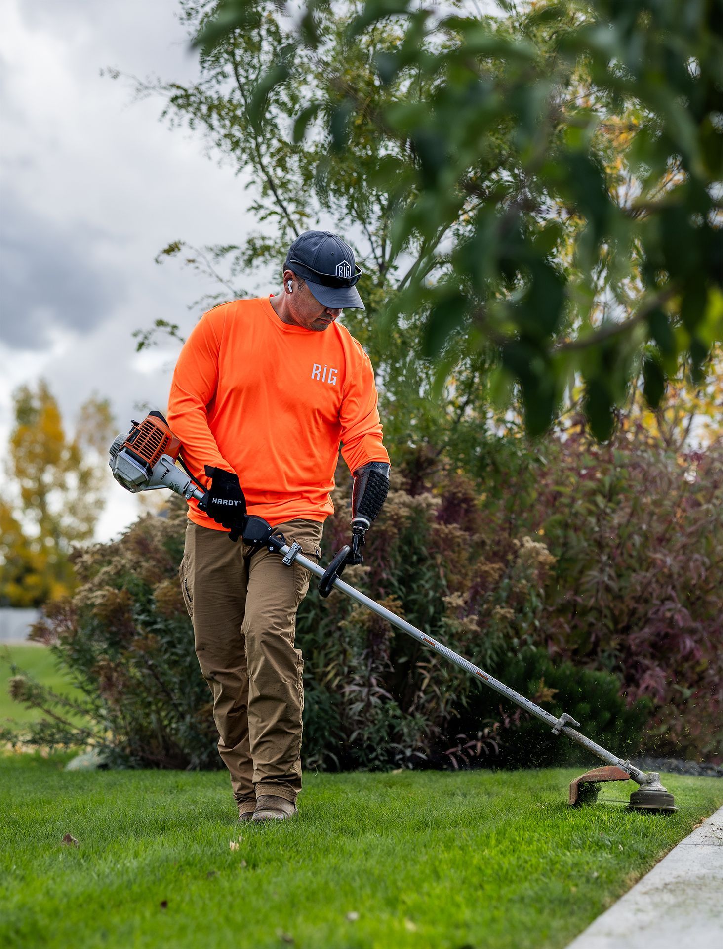 Man in orange shirt using a weed whacker on green grass next to a concrete walkway.