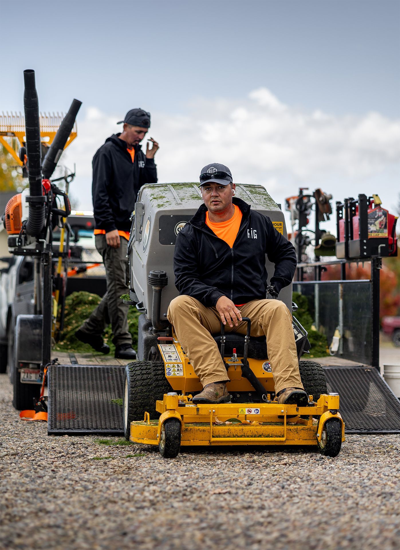 Two landscaping workers in black jackets, one seated on a yellow mower, the other near a trailer with equipment.