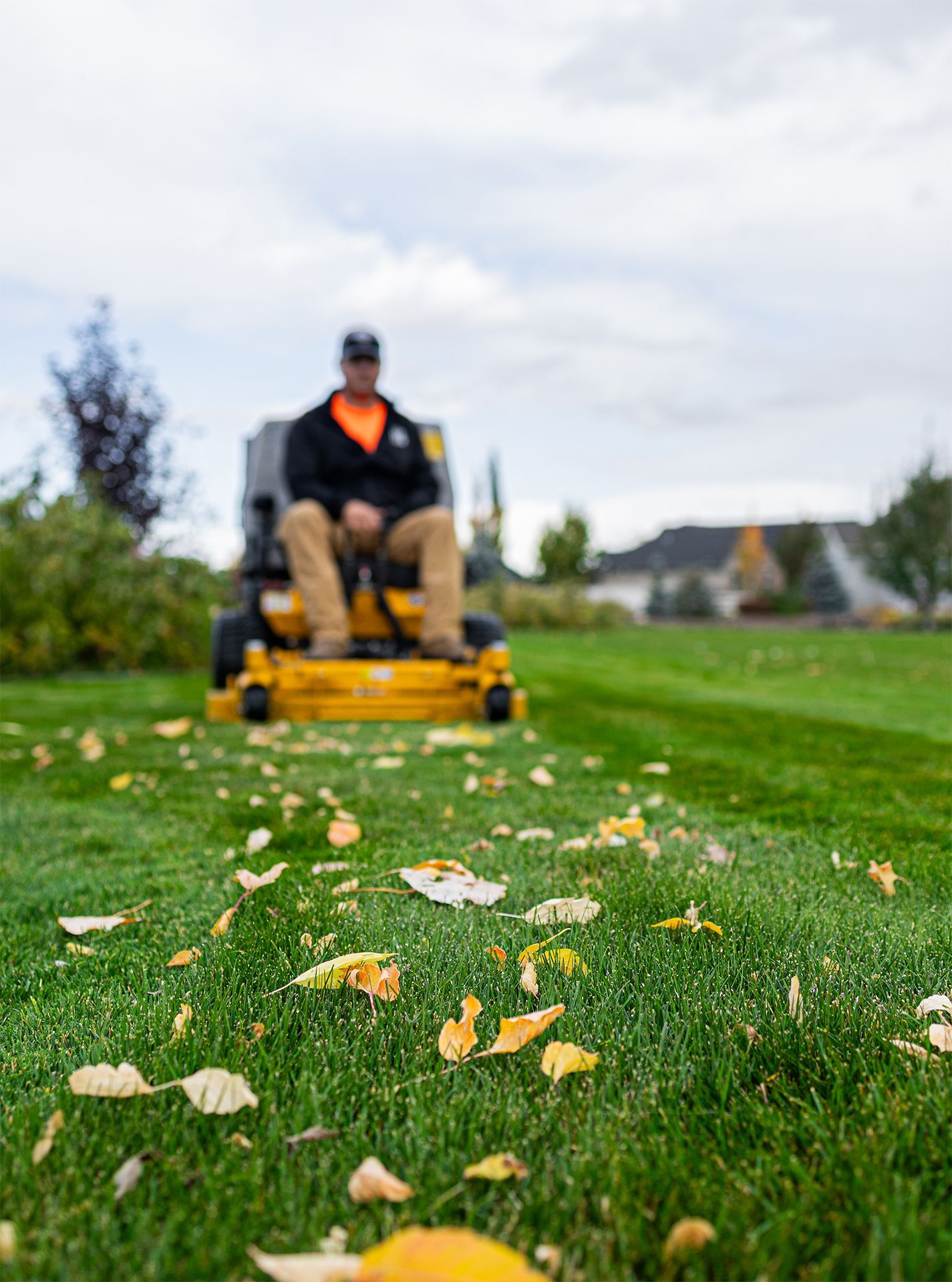 Person on a riding lawnmower cutting grass, with fallen leaves on the green lawn under a cloudy sky.