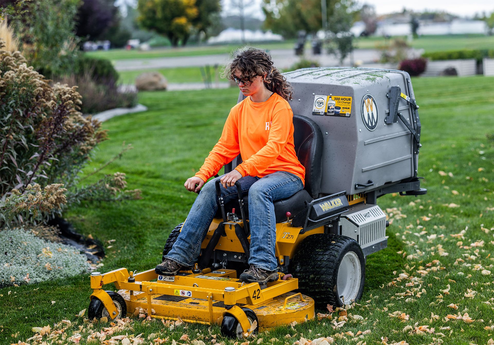 Woman in orange shirt operating a yellow riding lawnmower with a leaf collection system on a grassy lawn.