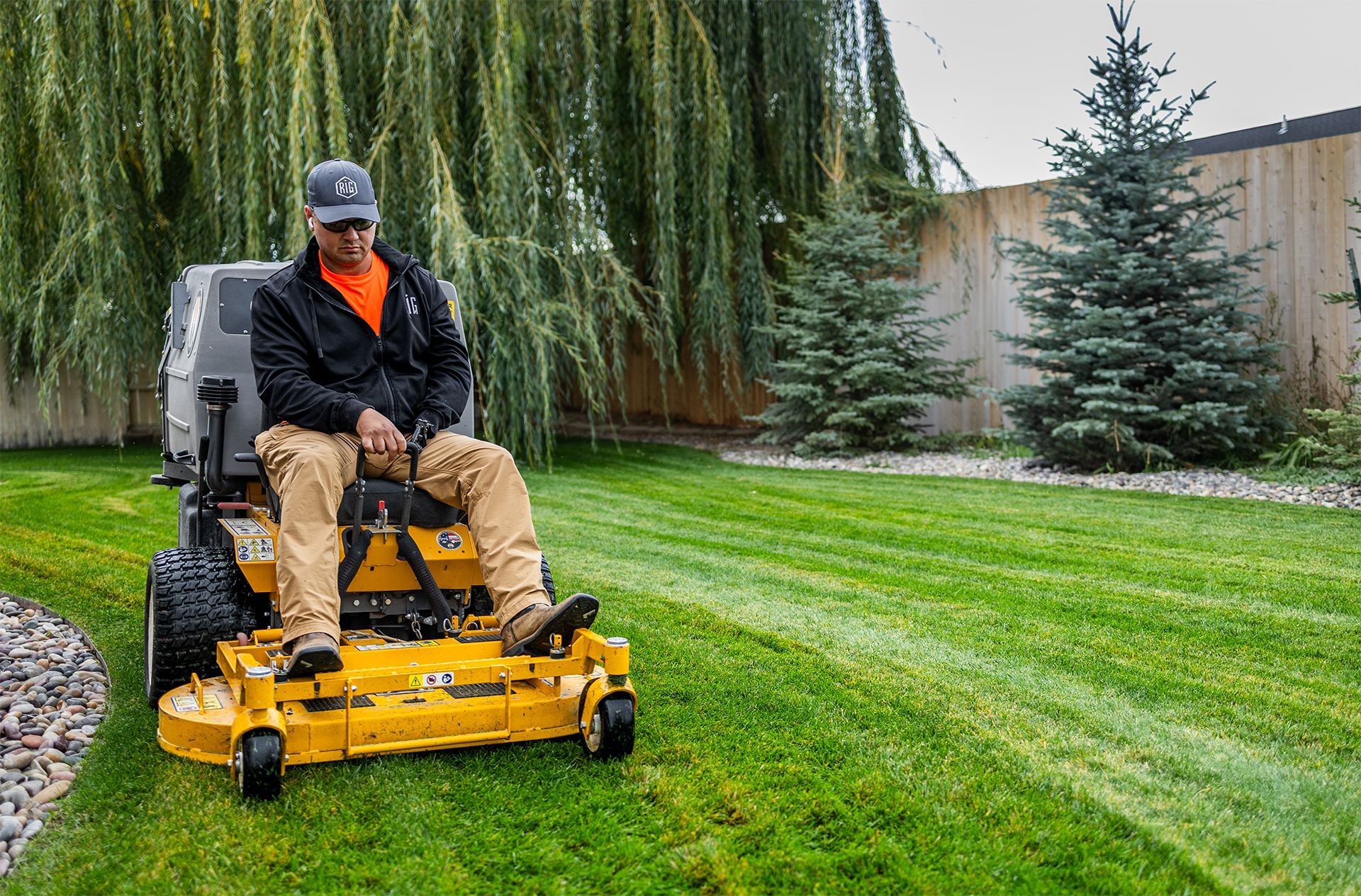 Man riding a yellow zero-turn lawnmower, mowing a green lawn near a house.