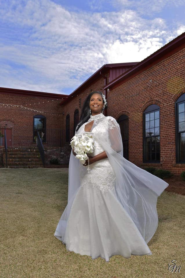 A bride in a white dress is standing in front of a brick building holding a bouquet of flowers.