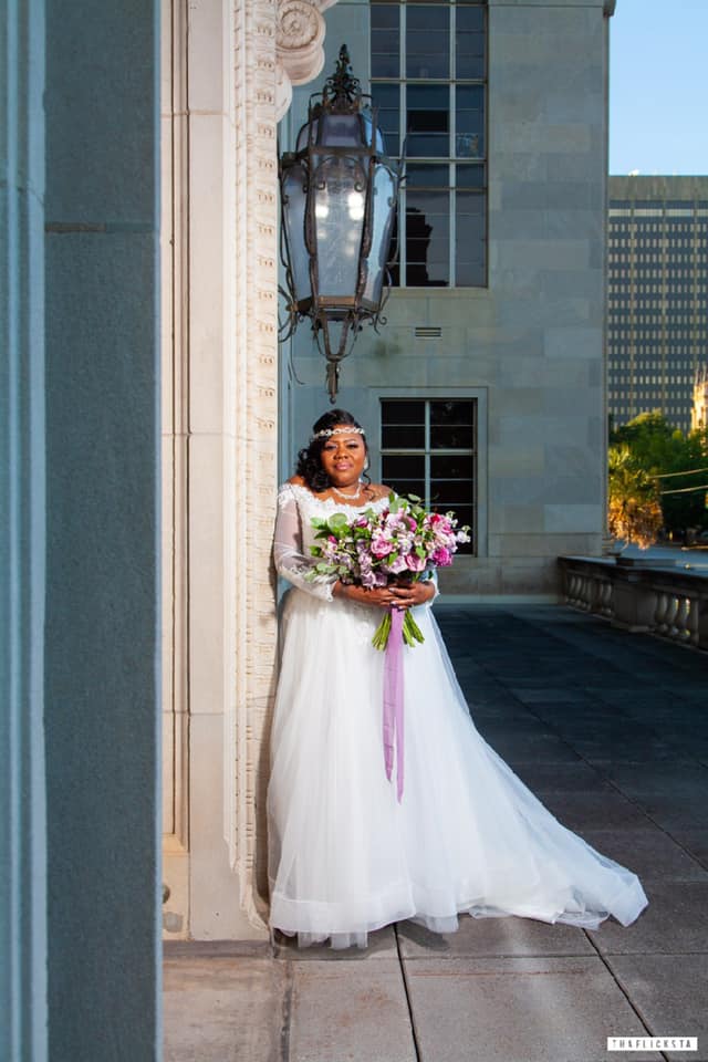 A bride in a wedding dress is standing next to a building holding a bouquet of flowers.