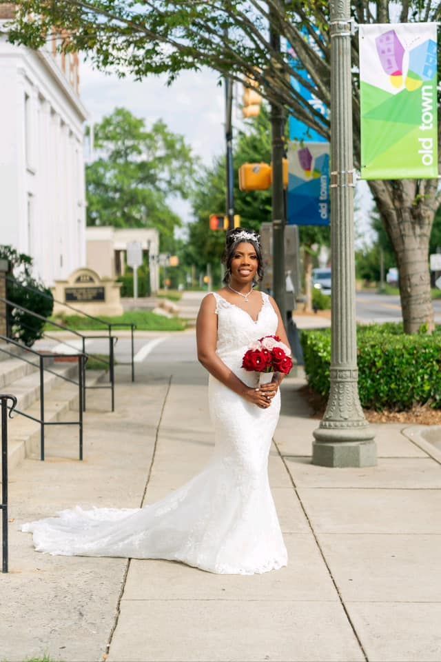 A bride in a white dress is standing on a sidewalk holding a bouquet of red roses.