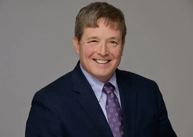 Man in navy suit, smiling, purple patterned tie, against a gray backdrop.
