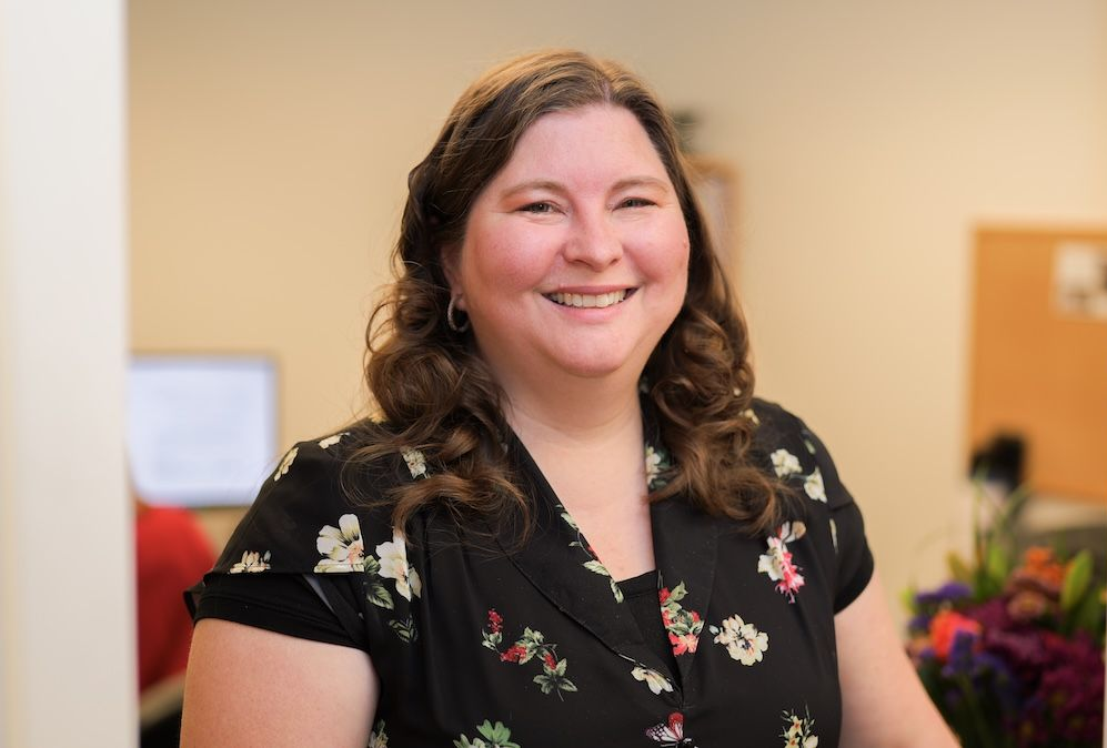 Woman with brown hair smiles in a black floral top, standing near a bulletin board and desk.