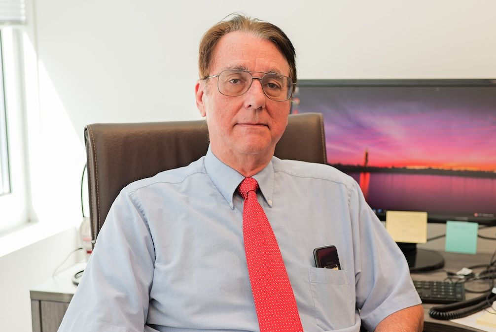 Man in glasses, red tie, seated at desk, looking at camera. Office setting with computer.