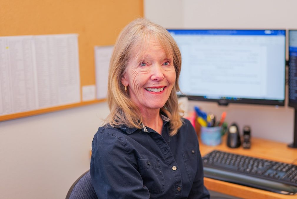 Woman smiling, sitting at a desk with two computer monitors, in an office.