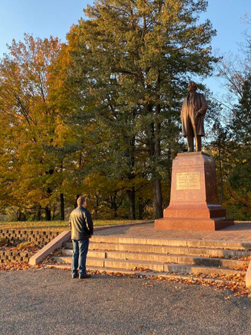 Three men in business attire pose outside a building, one with sunglasses, next to a blue art sculpture.