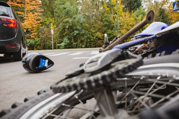 Motorcycle crashed at a crosswalk with a black car. A helmet lies on the ground, trees in the background.