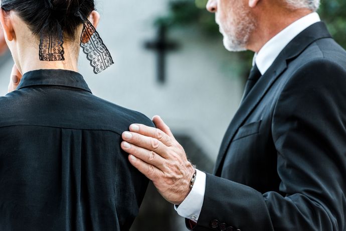 Man in suit comforts person in black clothing at a funeral, cross in the background.