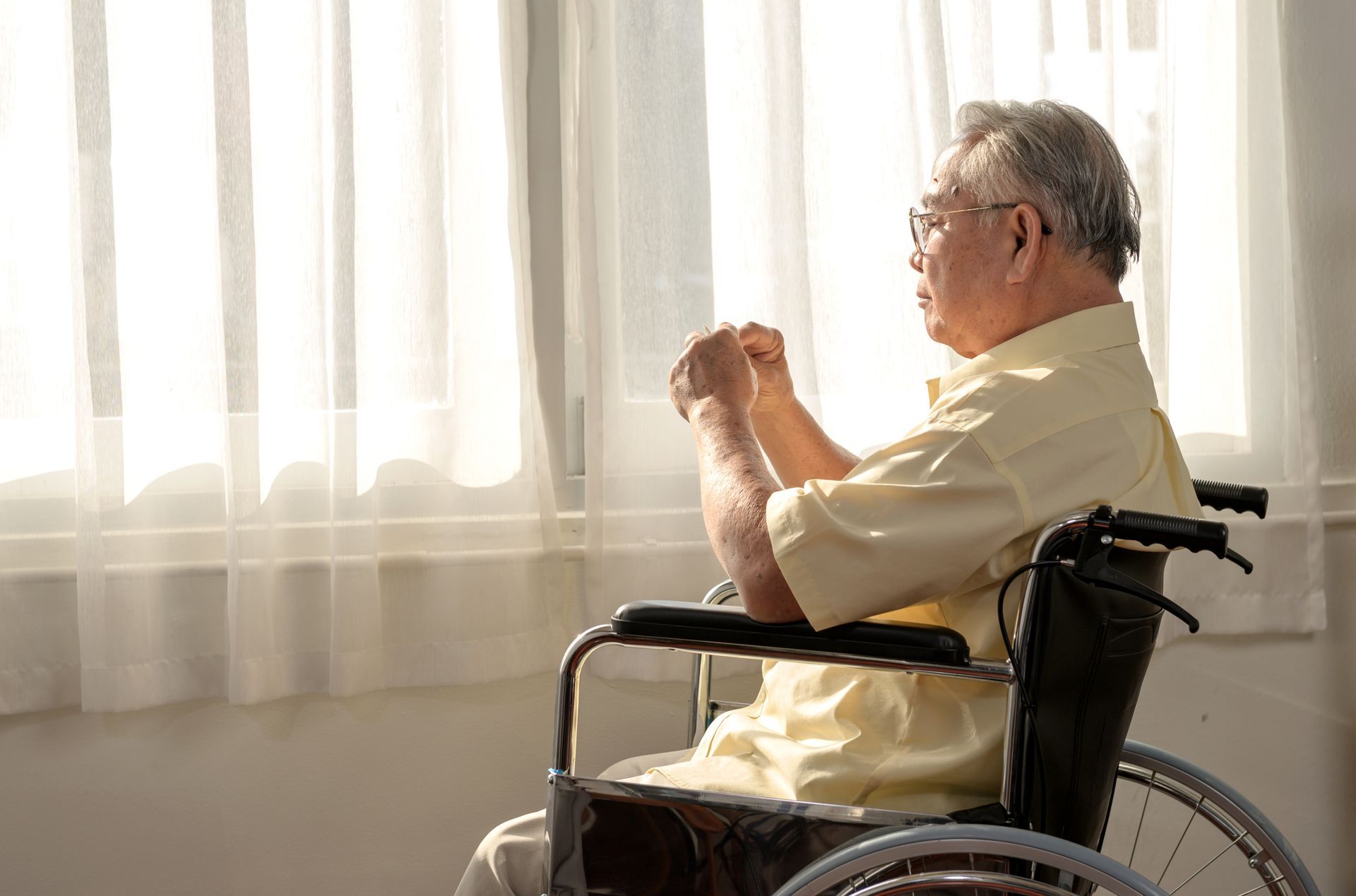 Older person in a wheelchair, looking out a window. Bright sunlight, hands clasped.