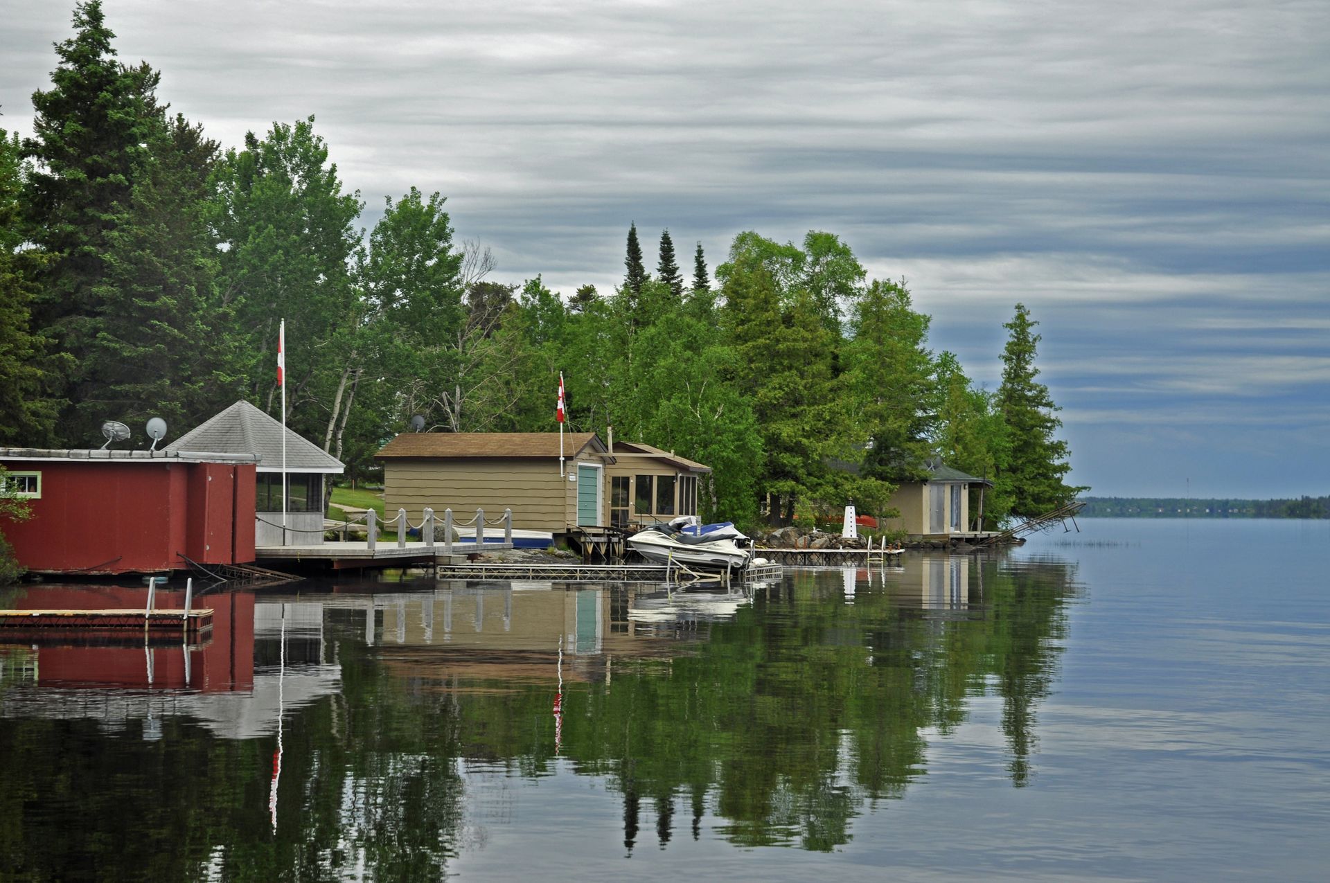 Lakeside cabins with boats reflected in calm water under a cloudy sky.