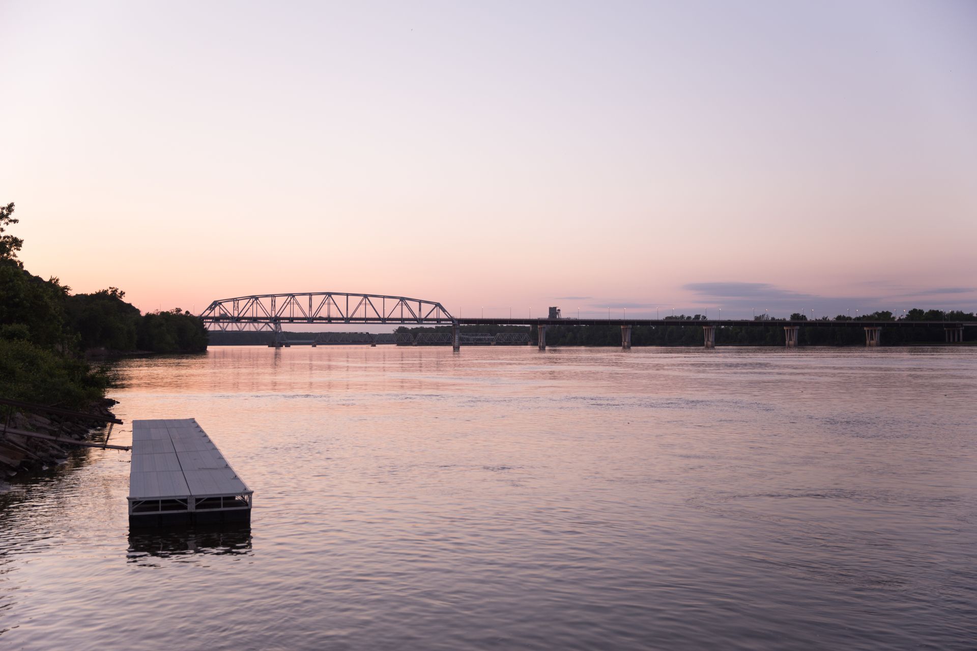 Sunset over a river with a bridge in the distance and a small dock in the foreground.