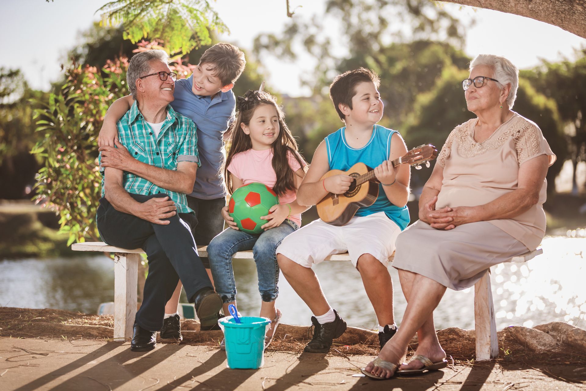 A family is sitting on a bench in a park and the boy is playing a small guitar for his grandma.